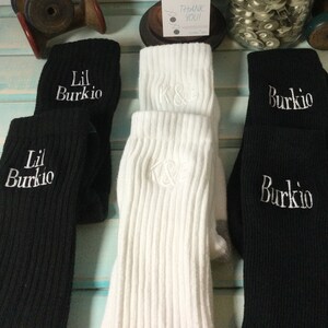 May include: A collection of ribbed knit socks in black and white. The black socks are embroidered with "Lil Burki0" and "Burkio" in white. The white socks are embroidered with "K&E" in white. The socks are folded over.