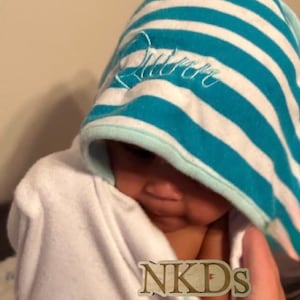 Puede incluir: Un beb&eacute; envuelto en una toalla con capucha blanca con rayas azules y blancas. La capucha tiene el nombre "Queen" bordado en escritura blanca. La cara del beb&eacute; es parcialmente visible. La imagen tiene el texto "NKDs" en la esquina inferior derecha.