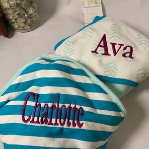 Puede incluir: Dos toallas con capucha personalizadas para beb&eacute;. Una toalla presenta rayas turquesas y blancas con el nombre "Charlotte" bordado en morado. La otra toalla es blanca con un patr&oacute;n de chevr&oacute;n turquesa y el nombre "Ava" bordado en morado.