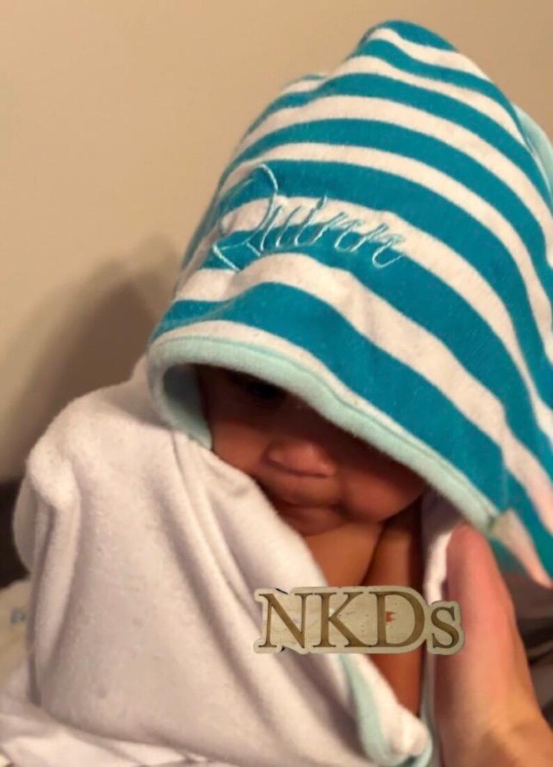 Puede incluir: Un beb&eacute; envuelto en una toalla con capucha blanca con rayas azules y blancas. La capucha tiene el nombre "Queen" bordado en escritura blanca. La cara del beb&eacute; es parcialmente visible. La imagen tiene el texto "NKDs" en la esquina inferior derecha.