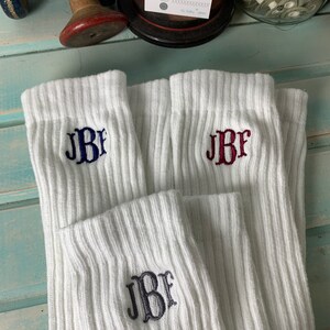 May include: White ribbed socks with embroidered initials in navy blue, burgundy, and charcoal gray. The socks are folded and displayed, showcasing the personalized monogram design. A small card with the text "THANK YOU!" is visible in the background.