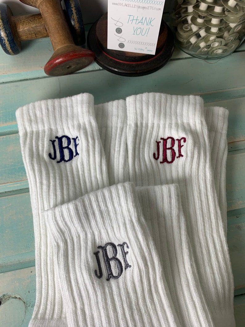 May include: White ribbed socks with embroidered initials in navy blue, burgundy, and charcoal gray. The socks are folded and displayed, showcasing the personalized monogram design. A small card with the text "THANK YOU!" is visible in the background.