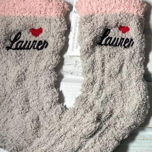 May include: A pair of gray fuzzy socks with a pink cuff. The name "Lauren" is embroidered in black script on each sock, topped with a small red heart. The socks are soft and cozy.