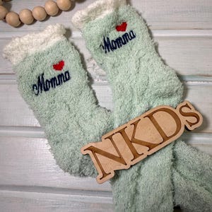 May include: A pair of light green fuzzy socks with white cuffs. The word "Momma" is embroidered in navy blue, with a red heart above the word. A wooden sign with the letters "NKDS" rests on the socks.