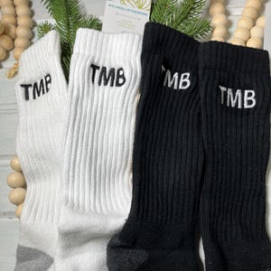 May include: A close-up shot of four ribbed crew socks. Two white socks and two black socks are displayed. Each sock has the embroidered letters "TMB" or "MB" in white. The socks are arranged with decorative wooden beads and greenery in the background.