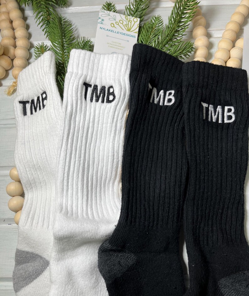 May include: A close-up shot of four ribbed crew socks. Two white socks and two black socks are displayed. Each sock has the embroidered letters "TMB" or "MB" in white. The socks are arranged with decorative wooden beads and greenery in the background.