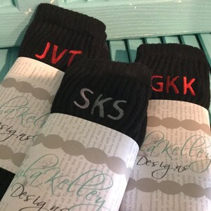 May include: Three pairs of black socks with embroidered initials in red and gray. Each pair is wrapped in paper with the text "la Kelley Designs". The initials are "JVT", "SKS", and "GKK".