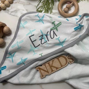 Puede incluir: Una manta de beb&eacute; blanca con un estampado de aviones azules y verdes. El nombre "Ezra" est&aacute; bordado en gris. La manta tiene un ribete gris. Juguetes de madera en el fondo.