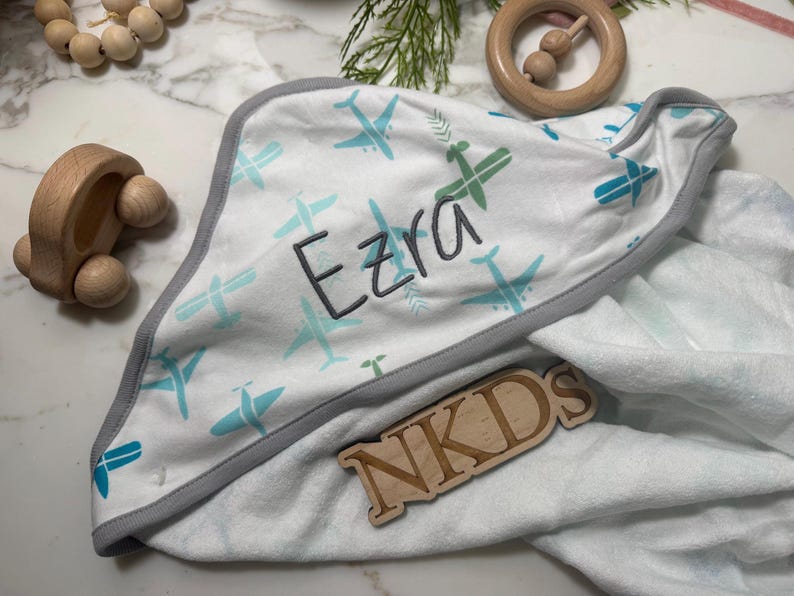 Puede incluir: Una manta de beb&eacute; blanca con un estampado de aviones azules y verdes. El nombre "Ezra" est&aacute; bordado en gris. La manta tiene un ribete gris. Juguetes de madera en el fondo.