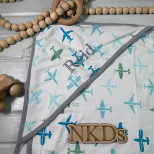 Puede incluir: Una manta blanca para beb&eacute; con un estampado de aviones azules y verdes. La manta tiene un borde gris y el nombre "Reid" bordado. Un avi&oacute;n de juguete de madera y una guirnalda de cuentas de madera tambi&eacute;n est&aacute;n en la imagen.
