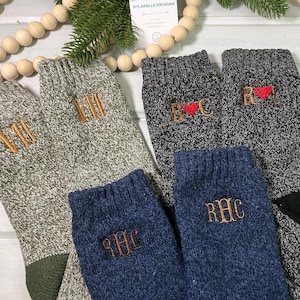 May include: Assorted pairs of cozy knit socks in gray, navy, and black. Each sock features embroidered initials in gold thread. Some socks have a heart symbol. A wooden bead garland and greenery are in the background.