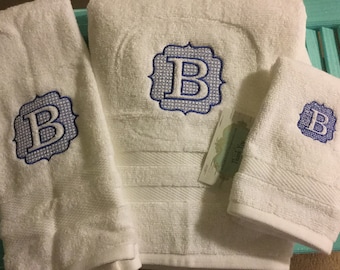 May include: Three white towels with blue embroidered letters "B" in a decorative frame. The towels are folded and arranged on a light blue surface.