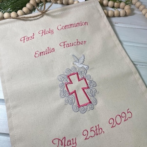 May include: A white canvas banner with pink embroidery that reads "First Holy Communion Emilia Faucher May 25th, 2025". The banner also features a white dove with a red cross in a gray swirl design.