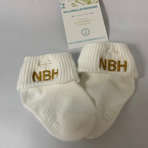 May include: Two white baby socks with gold embroidered cross and initials "NBH".