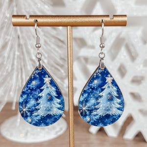 May include: A pair of teardrop-shaped earrings with a blue and white watercolor design of a Christmas tree with snowflakes. The earrings are made of wood and have silver hooks.