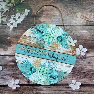 Pu&ograve; includere: Un cartello rotondo in legno con uno sfondo in legno rovinato blu e bianco. Il cartello presenta un disegno floreale con fiori blu-verdi e bianchi e dettagli dorati. Il testo "The D'Abraccio's" &egrave; scritto in nero su una striscia blu.