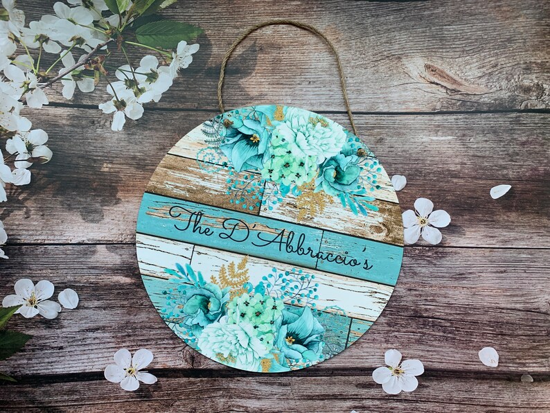 Pu&ograve; includere: Un cartello rotondo in legno con uno sfondo in legno rovinato blu e bianco. Il cartello presenta un disegno floreale con fiori blu-verdi e bianchi e dettagli dorati. Il testo "The D'Abraccio's" &egrave; scritto in nero su una striscia blu.