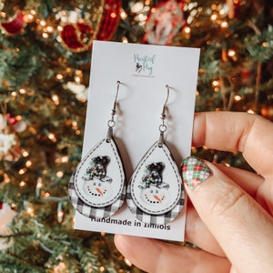 May include: A pair of dangle earrings featuring a snowman design with a black top hat, white face, and black eyes. The earrings are made of wood and have a black and white checkered pattern. The earrings are handmade in Illinois. The earrings are on a white card with the text "Painted Pug" in black.
