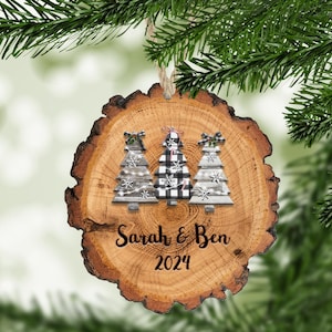 May include: A wooden Christmas ornament with a rustic wood slice design. Three small Christmas trees are decorated with black and white buffalo plaid and snowflakes. The ornament is personalized with the names "Sarah & Ben" and the year "2024".