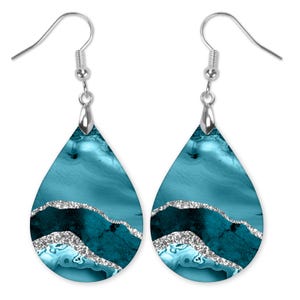 May include: Teal and silver teardrop shaped dangle earrings with a silver hook. The earrings have a marbled pattern with silver glitter accents.