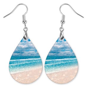 May include: A pair of teardrop-shaped earrings with a beach scene printed on them. The earrings feature a blue sky with white clouds, a blue ocean with white waves, and a sandy beach.