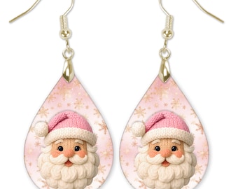 Può includere: Un paio di orecchini pendenti a goccia con la grafica di un volto di Babbo Natale con un berretto di lana rosa e una barba bianca. Lo sfondo è rosa tenue con motivi a fiocchi di neve dorati. Gli orecchini hanno ganci in metallo dorato.