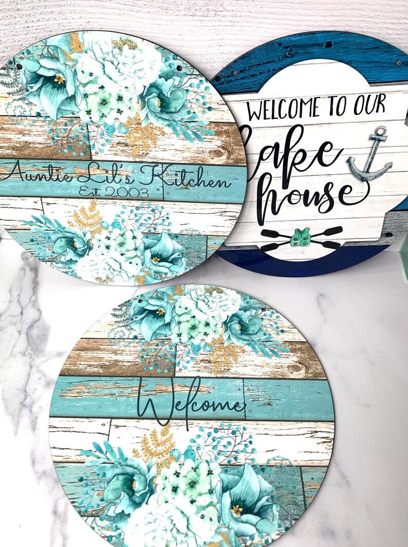 Pu&ograve; includere: Tre cartelli rotondi in legno con sfondo a venature di legno blu e bianco e disegni floreali. I cartelli recitano "Auntie Lil's Kitchen Est 2003", "Benvenuti alla nostra casa sul lago" con un'ancora e "Benvenuti".