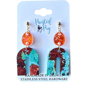 May include: Dangle earrings featuring a round orange glitter stud connected to a larger arch-shaped pendant with a mottled turquoise and dark brown pattern. The earrings are displayed on a white card with the text Painted Pug and Stainless Steel Hardware.