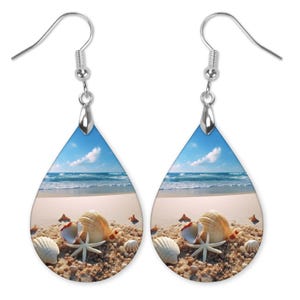 May include: A pair of teardrop-shaped earrings with a beach scene and seashells. The earrings are silver and have a blue sky, white sand, and ocean waves in the background. The seashells are white and brown and are arranged in a cluster on the sand.