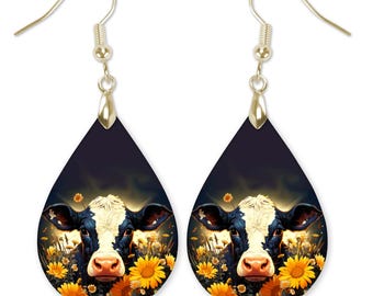 Puede incluir: Un par de pendientes en forma de lágrima con fondo negro y una imagen colorida de la cara de una vaca rodeada de flores amarillas.