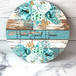 Pu&ograve; includere: Segno rotondo in legno con uno sfondo in legno invecchiato blu e marrone. Il segno presenta un motivo floreale con fiori bianchi e turchesi e accenti di glitter dorati. Il testo "Home Sweet Home" &egrave; scritto in lettere corsive nere.