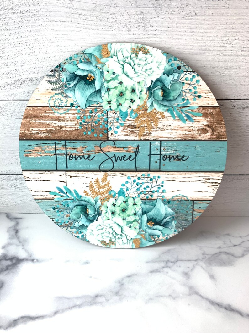 Pu&ograve; includere: Segno rotondo in legno con uno sfondo in legno invecchiato blu e marrone. Il segno presenta un motivo floreale con fiori bianchi e turchesi e accenti di glitter dorati. Il testo "Home Sweet Home" &egrave; scritto in lettere corsive nere.