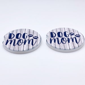 May include: Two white ceramic coasters with a woodgrain pattern. The coasters feature the text "Dog Mom" in blue with paw prints.