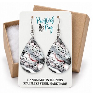 May include: Teardrop-shaped earrings featuring a baseball pattern with red stitching on a black, white, and silver glitter background. The display card reads Painted Pug, Handmade in Illinois, and Stainless Steel Hardware.