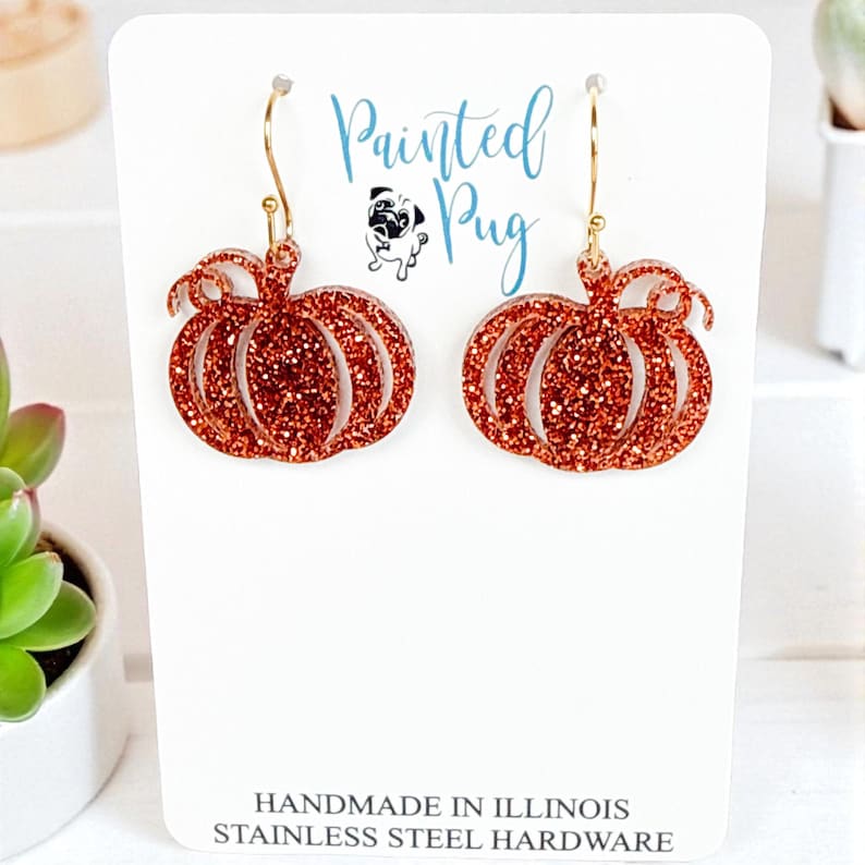 May include: A pair of orange glitter pumpkin-shaped dangle earrings with gold-tone hooks. The display card features the text Painted Pug, a pug illustration, and the text HANDMADE IN ILLINOIS STAINLESS STEEL HARDWARE at the bottom.
