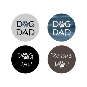 May include: Four round stickers with different backgrounds and text. The stickers say "Dog Dad" with a paw print in the middle of the "D" and "G". One sticker is gray, one is blue, one is black, and one is brown. The brown sticker says "Rescue Dad" with a paw print in the middle of the "D".