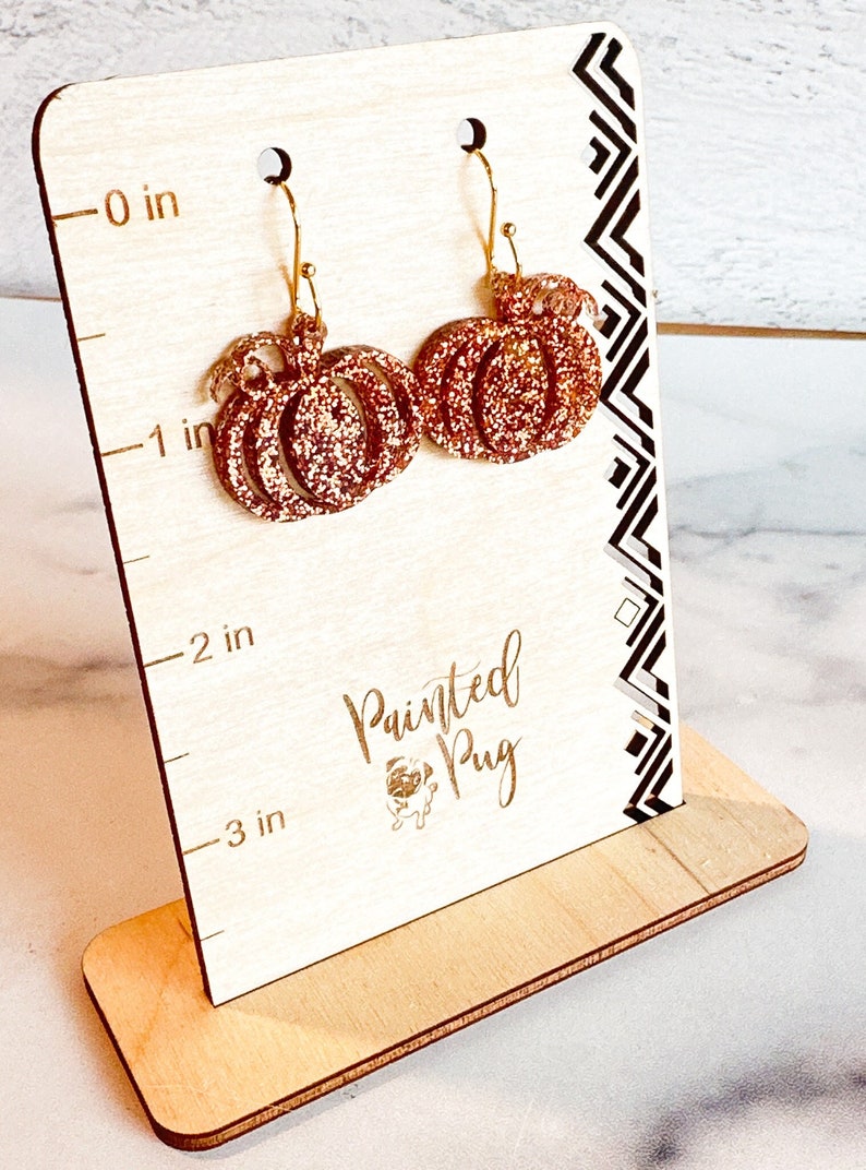May include: A pair of copper-colored glitter pumpkin earrings with gold-tone hooks, displayed on a light wood stand featuring a ruler marked in inches and the text Painted Pug with a small pug icon, measuring approximately 1.5 inches in height.