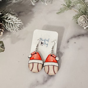 May include: A pair of dangle earrings featuring a baseball with a Santa hat on top. The earrings are painted in red, white, and brown colors. The earrings are on a white card with the text "Painted Pug" on it.