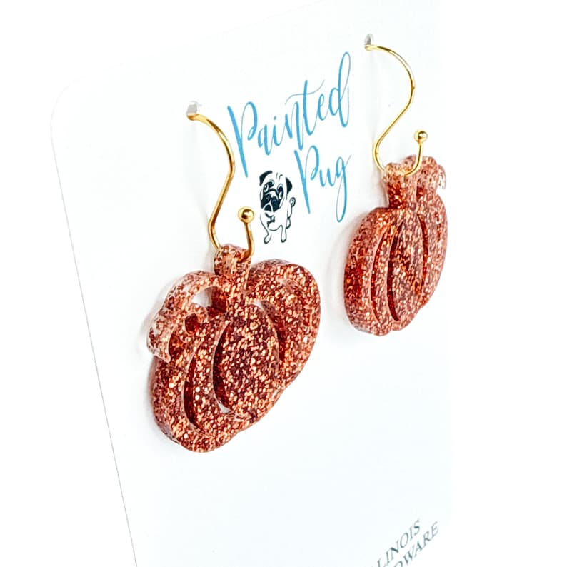 May include: A pair of dangling earrings featuring orange glitter pumpkin-shaped charms attached to gold-toned curved ear wires. The earrings are displayed on a white card with the text Painted Pug and a small illustration of a pug dog.