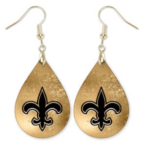 May include: A pair of gold teardrop-shaped earrings with a black fleur-de-lis design. The earrings have a sparkly gold finish.