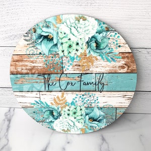 Pu&ograve; includere: Un cartello rotondo in legno con uno sfondo in legno invecchiato e un design floreale blu-verde. Il cartello dice "The Cox Family" in lettere nere.
