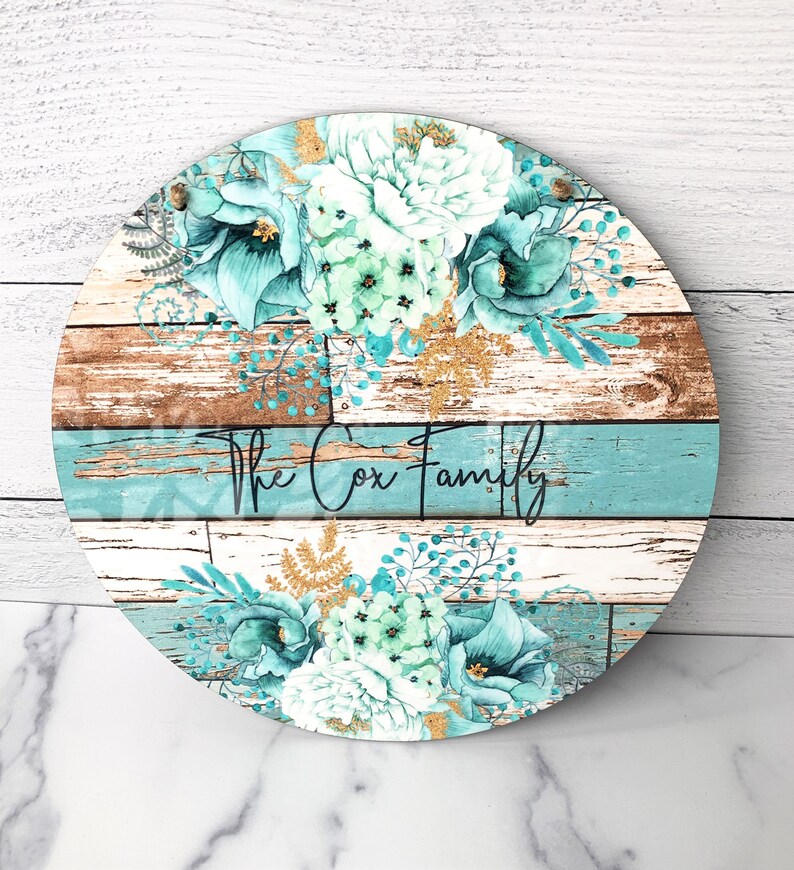 Pu&ograve; includere: Un cartello rotondo in legno con uno sfondo in legno invecchiato e un design floreale blu-verde. Il cartello dice "The Cox Family" in lettere nere.