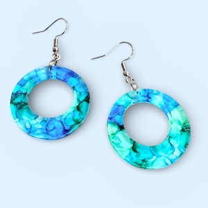 May include: A pair of hoop earrings with a marbled design. The earrings feature a silver hook and a circular hoop with shades of blue, teal, and green. The marbled effect gives the earrings a unique, artistic look.