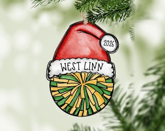 May include: A flat, illustrated Christmas ornament shaped like a cheerleader pom-pom wearing a red Santa hat with a white brim. The text "WEST LINN" is printed on the white brim, and the number "2026" is on the hat's white pom-pom.
