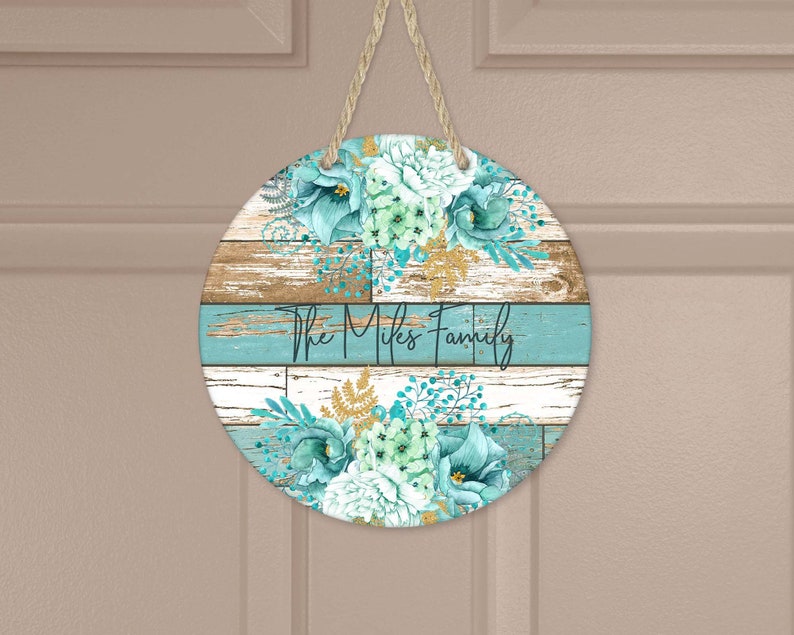 Pu&ograve; includere: Un cartello rotondo in legno con una finitura verniciata invecchiata blu e bianca. Il cartello presenta un motivo floreale con fiori blu-verdi e bianchi e dettagli dorati. Il testo "The Mikes Family" &egrave; scritto in nero sul cartello.