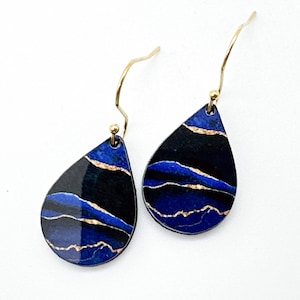 May include: A pair of teardrop-shaped earrings with a blue and black marbled design and gold accents. The earrings have a gold hook closure.