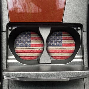 May include: Two round American flag themed car coasters with a distressed wood look. The coasters are in a car's cup holder.