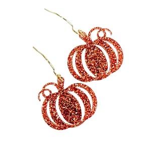 May include: A pair of dangle earrings featuring pumpkin-shaped charms covered in orange glitter. Each pumpkin charm has a small curled stem at the top and is attached to a thin gold-toned metal ear wire hook for hanging from the earlobe.