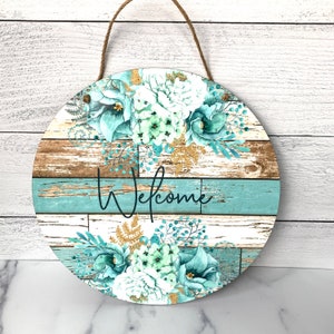 Pu&ograve; includere: Un cartello rotondo in legno con uno sfondo in legno invecchiato e un design floreale blu-verde e bianco. Il cartello dice "Benvenuti" in scrittura nera.