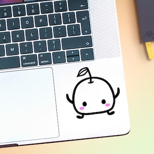 Puede incluir: Una pegatina en blanco y negro de una manzana de dibujos animados con un tallo y una hoja. La manzana tiene una cara linda con mejillas rosadas y una pequeña sonrisa. La pegatina está en una computadora portátil plateada.